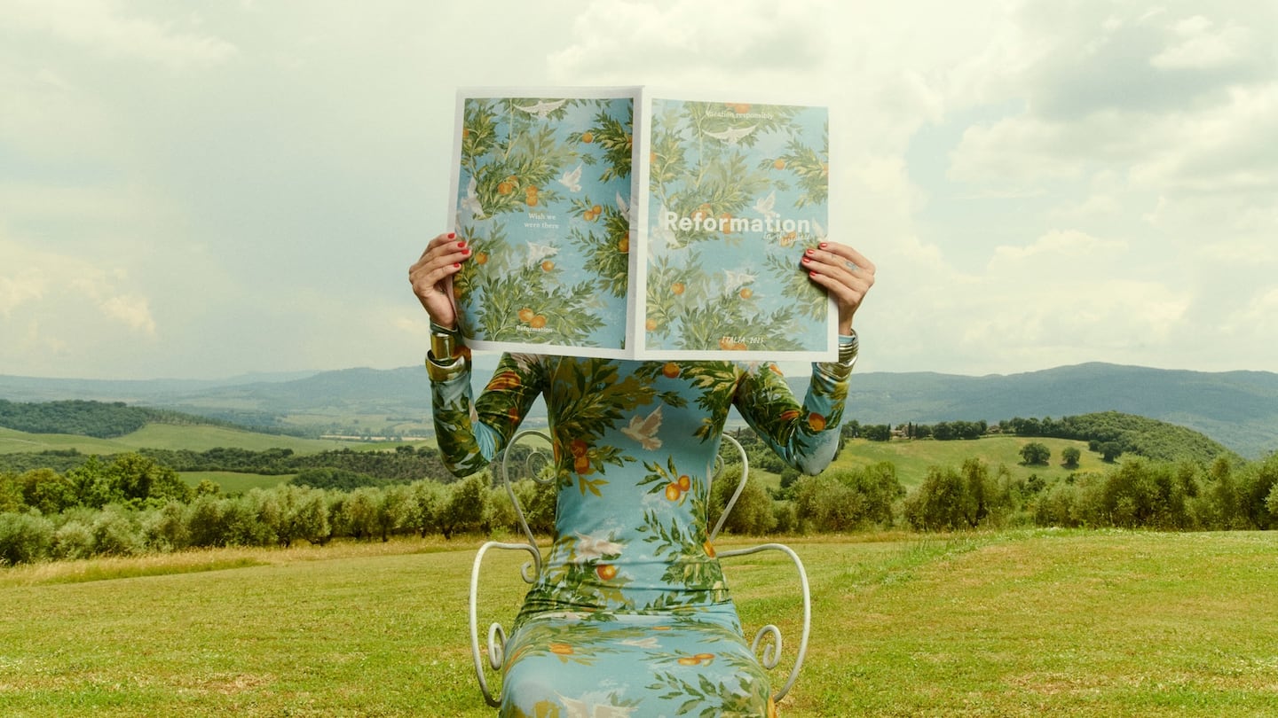 A model wears a long, botanical-patterned Reformation dress while sitting on a white chair in a scenic grassy landscape, holding up a newspaper with a matching floral print.